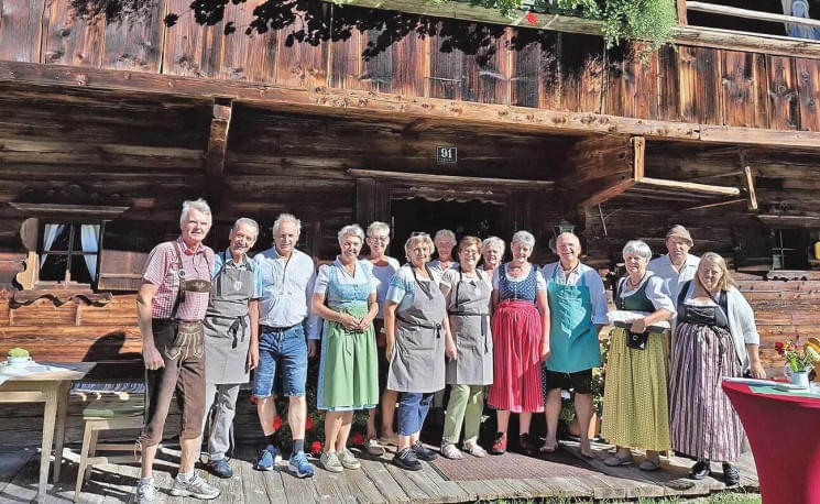 Eine Gruppe von älteren Menschen in traditioneller Kleidung posiert auf einer Holzterrasse vor einem rustikalen Gebäude bei sonnigem Wetter.