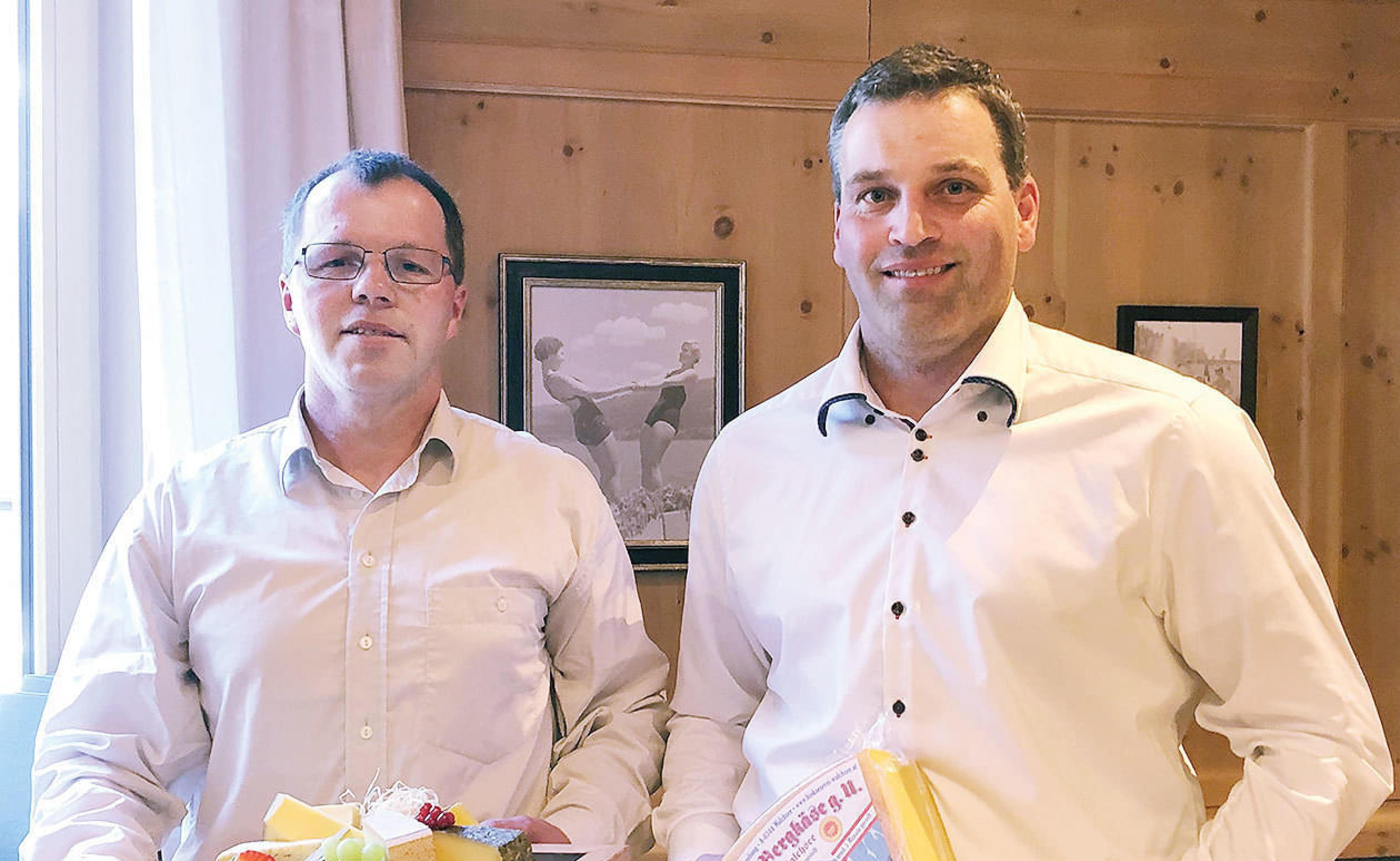 Zwei Männer in weißen Hemden stehen lächelnd nebeneinander in einem Raum mit Holzwänden. Einer hält einen Präsentkorb in den Händen.