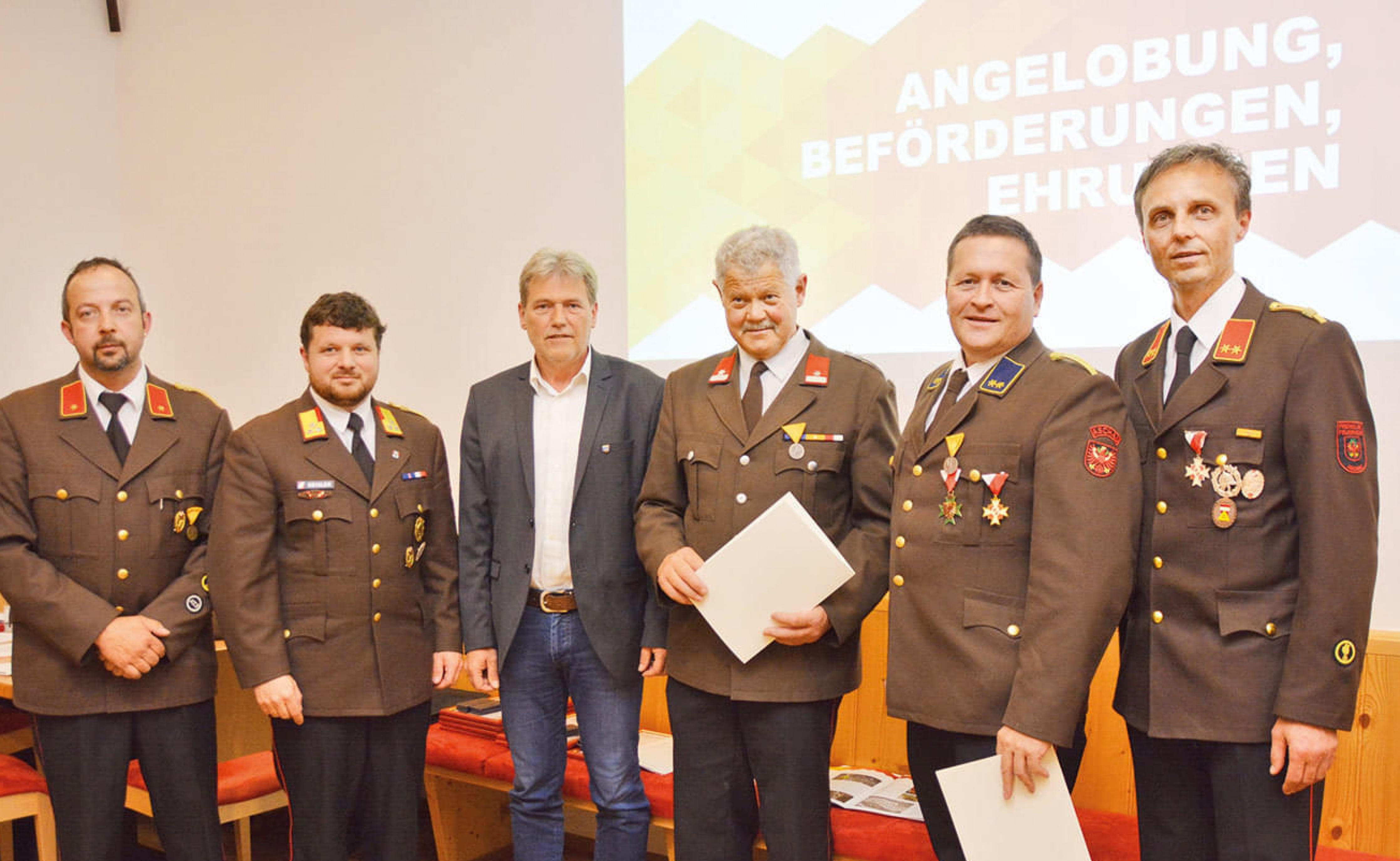 Gruppe von Männern in Uniformen bei einer Zeremonie mit Auszeichnungen. Eine Leinwand im Hintergrund zeigt Texte zur Ehrung und Beförderung.