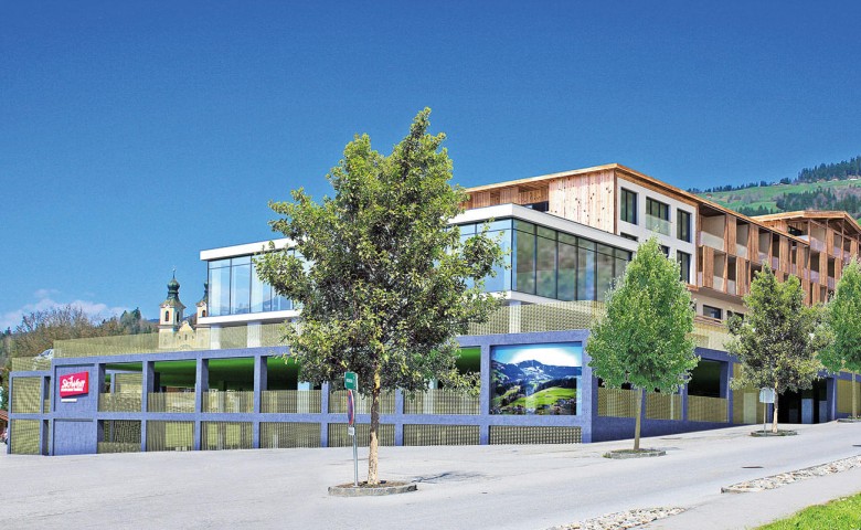 Modernes Holzgebäude mit großen Fenstern und grünem Zaun, umgeben von Bäumen. Im Hintergrund sind Berge und ein strahlend blauer Himmel zu sehen.