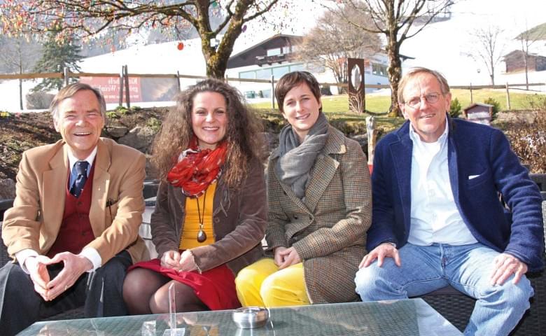 Vier Erwachsene sitzen draußen an einem Tisch in einer ländlichen Umgebung und lächeln in die Kamera. Im Hintergrund sind Bäume und ein Gebäude zu sehen.