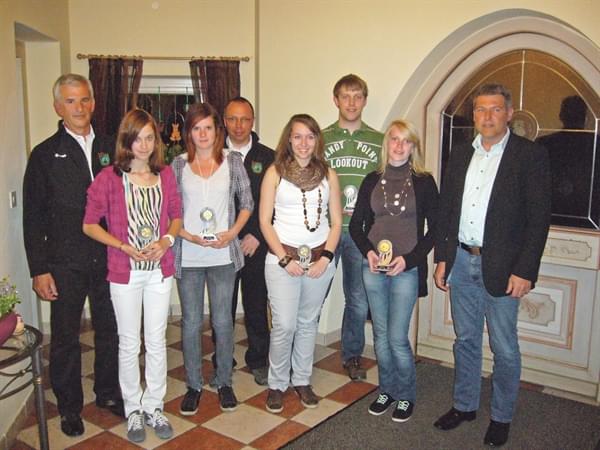 Gruppe von acht Personen steht in einem Raum mit Fliesenboden und einem großen Spiegel, drei davon halten Trophäen, alle sind lässig bis formell gekleidet.