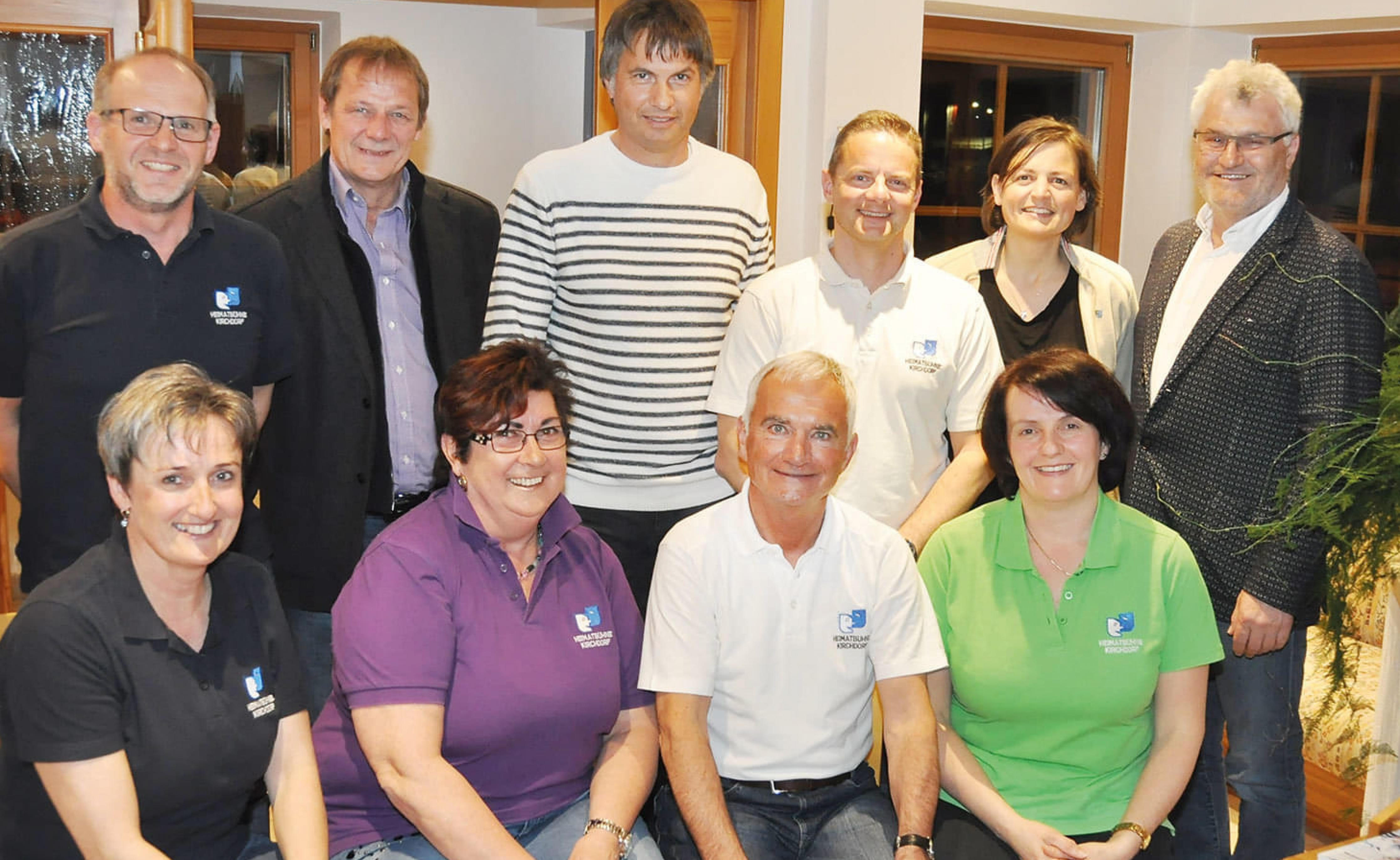 Eine Gruppe von zehn Menschen, fünf sitzend und fünf stehend, lächelt in die Kamera in einem Raum mit warmen Farben und Holzfenstern.