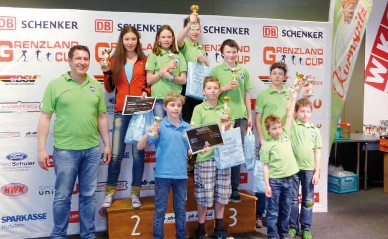 Gruppe von Kindern und Jugendlichen in grünen T-Shirts posiert mit Pokalen auf einem Siegerpodest bei einem Wettbewerb, Hintergrund mit Sponsorenlogos.