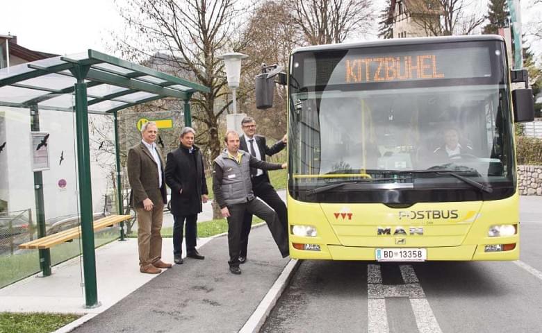 Vier Männer posieren neben einem gelben Bus mit der Aufschrift "Kitzbühel" an einer Bushaltestelle. Der Busfahrer steht in der Tür.