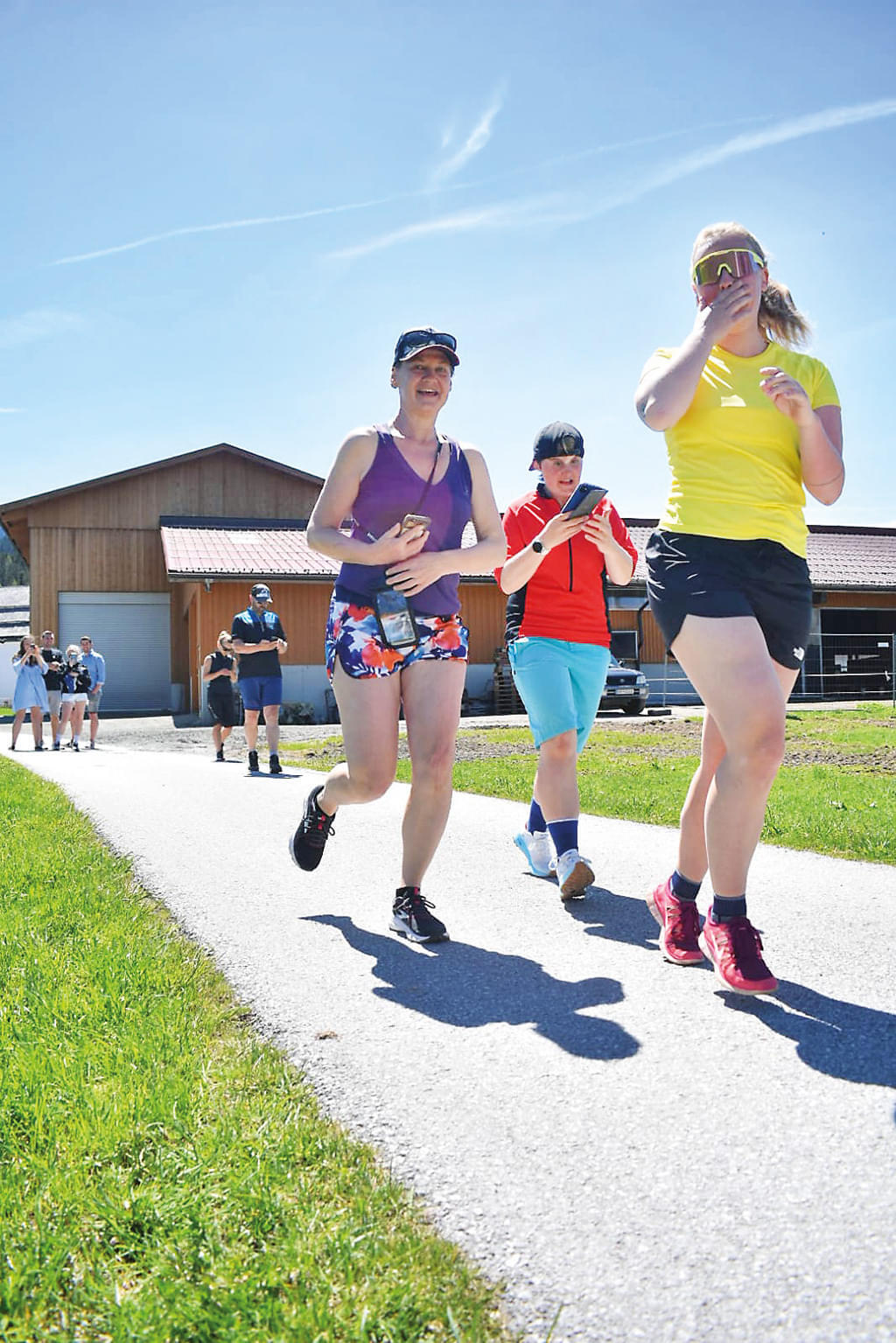 Mehrere Personen joggen auf einem sonnigen Weg, gesäumt von grünen Wiesen und einem Holzhäuschen im Hintergrund. Alle tragen sportliche Kleidung.