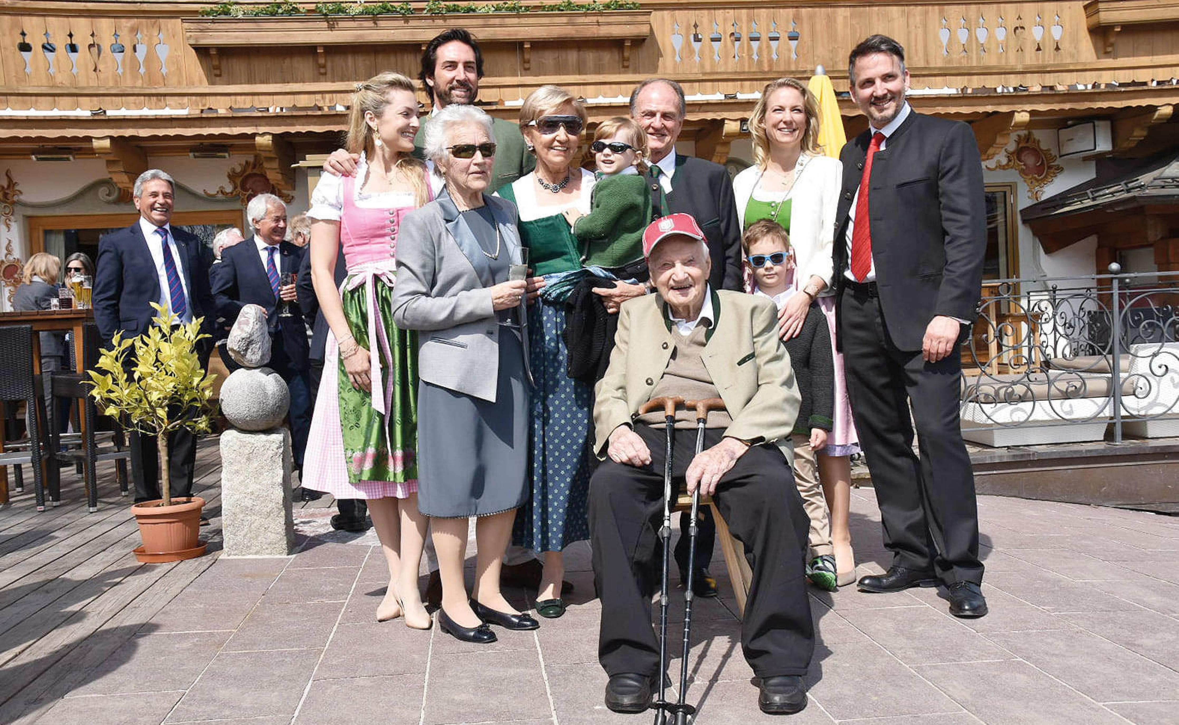 Eine Gruppe von Menschen, darunter ein älterer Mann mit Stock, posiert lächelnd im Freien vor einem traditionellen Gebäude mit Holzbalkon.