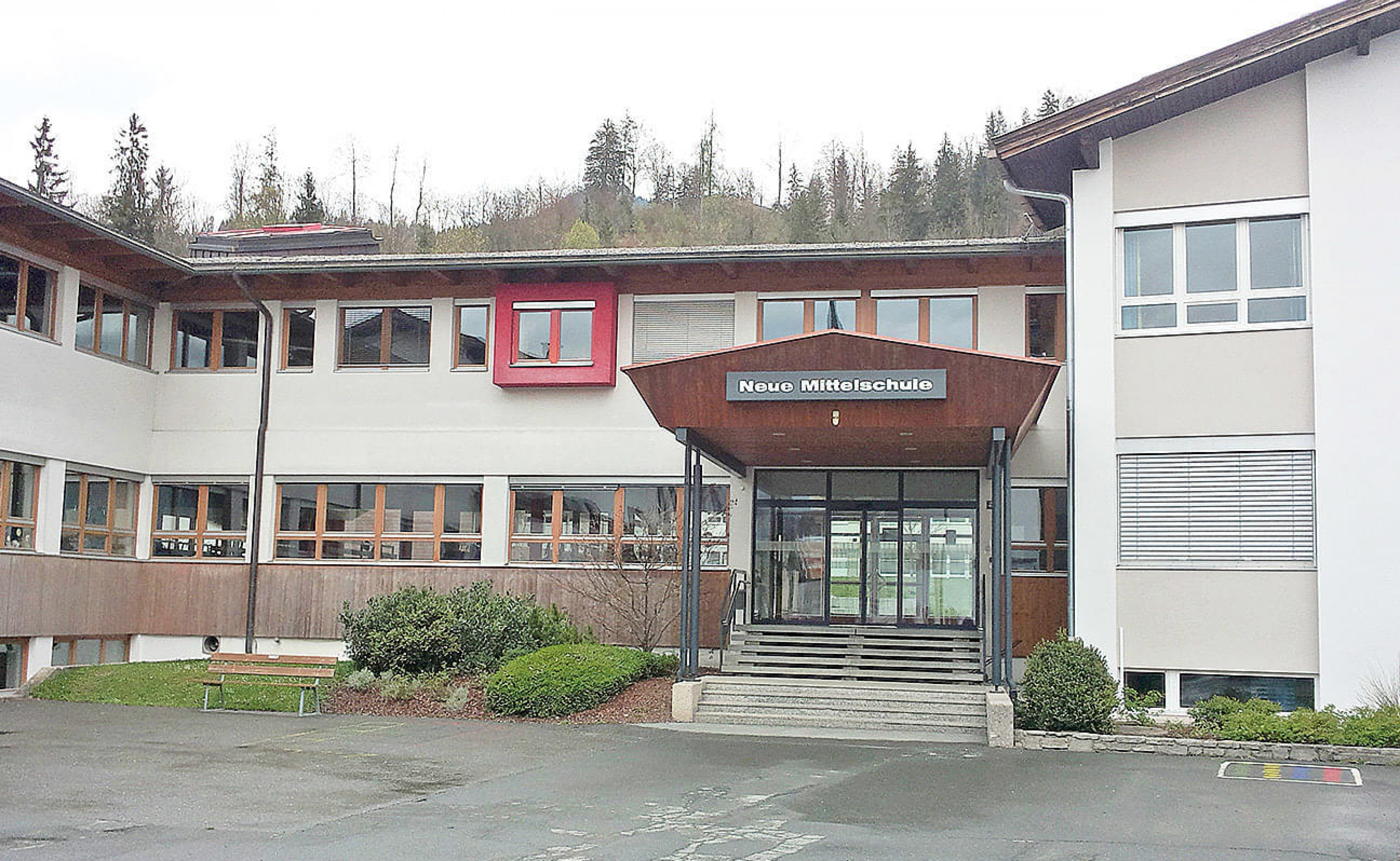 Zweistöckiges Schulgebäude mit großen Fenstern und einem überdachten Eingangsbereich, umgeben von Bäumen und einem Parkplatz im Vordergrund.