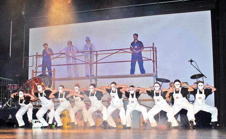 Eine Gruppe von Tänzern in weißen Overalls und schwarz-weißer Gesichtsbemalung führt eine Choreografie auf einer Bühne mit Malergerüsten im Hintergrund auf.