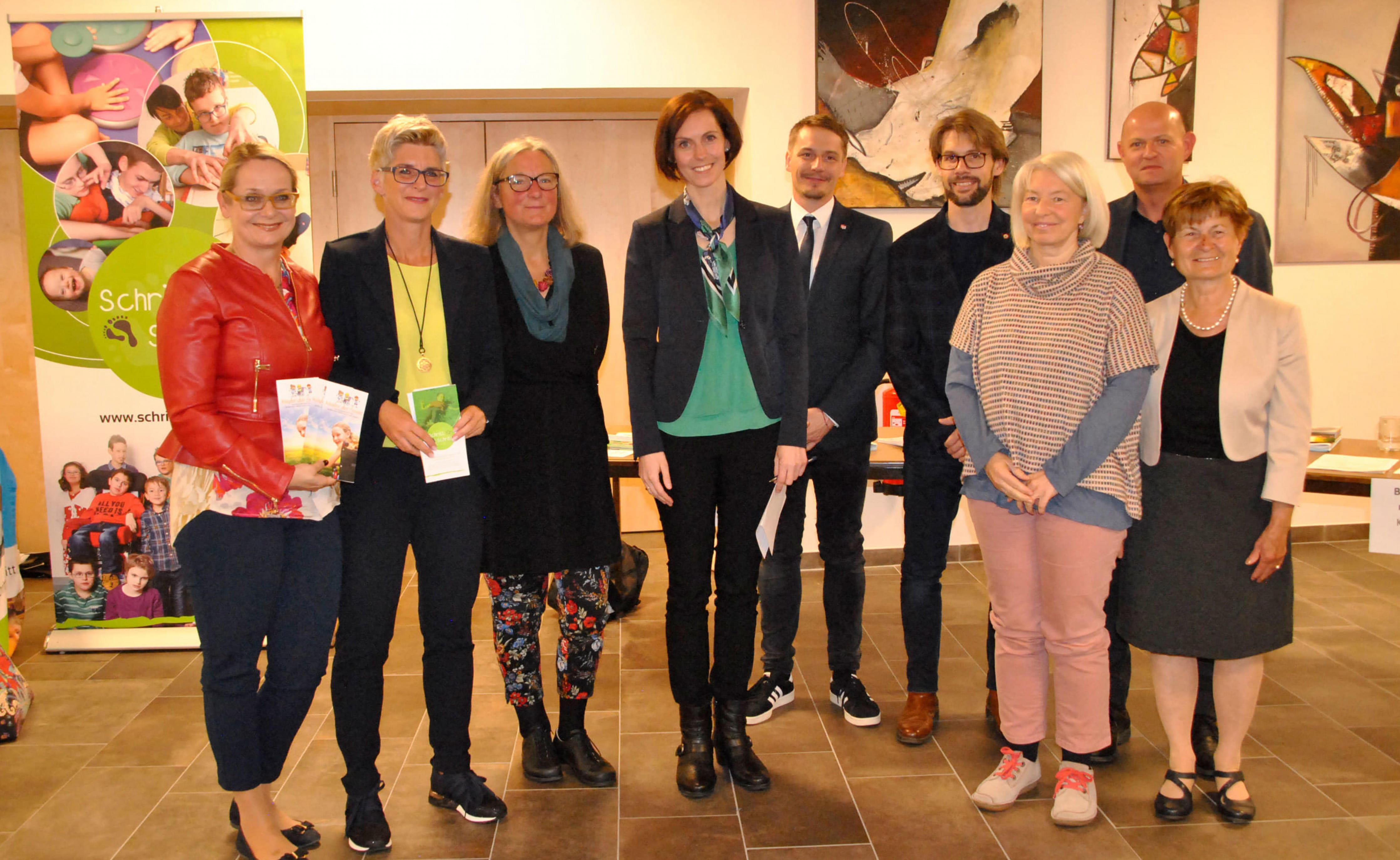 Eine Gruppe von zehn Personen steht lächelnd zusammen in einem Raum mit Kunstwerken an der Wand. Einige halten Bücher oder Broschüren in den Händen.