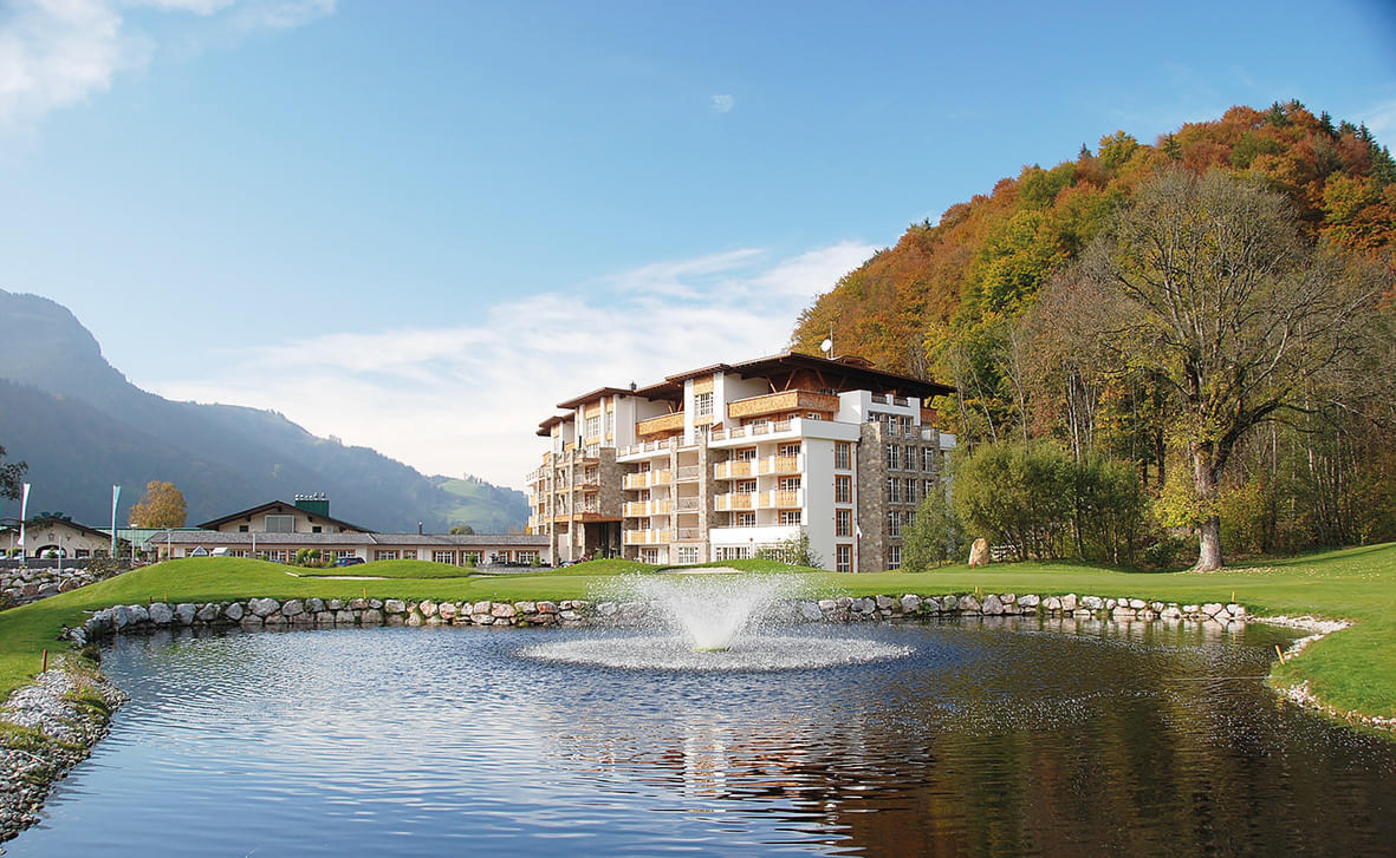 Großes Hotelgebäude am Ufer eines Teichs mit Springbrunnen, umgeben von Hügeln und buntem Herbstlaub unter blauem Himmel.