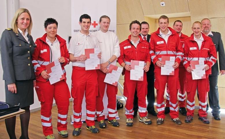 Eine Gruppe von Personen in Rotkreuz-Uniformen steht lächelnd mit Zertifikaten. Daneben steht eine Frau in einer anderen Uniform.