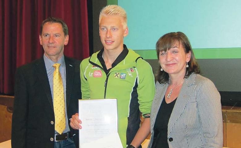 Drei Personen stehen nebeneinander, die mittlere Person hält ein Papier. Eine Frau und zwei Männer lächeln in die Kamera. Hintergrund mit rotem Vorhang.