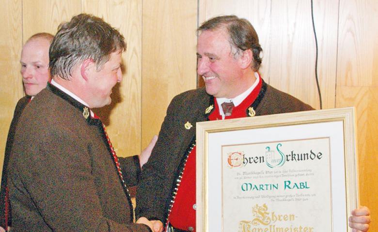 Zwei Männer in traditioneller Tracht schütteln Hände und halten eine Urkunde. Beide lächeln, während ein dritter Mann im Hintergrund steht.