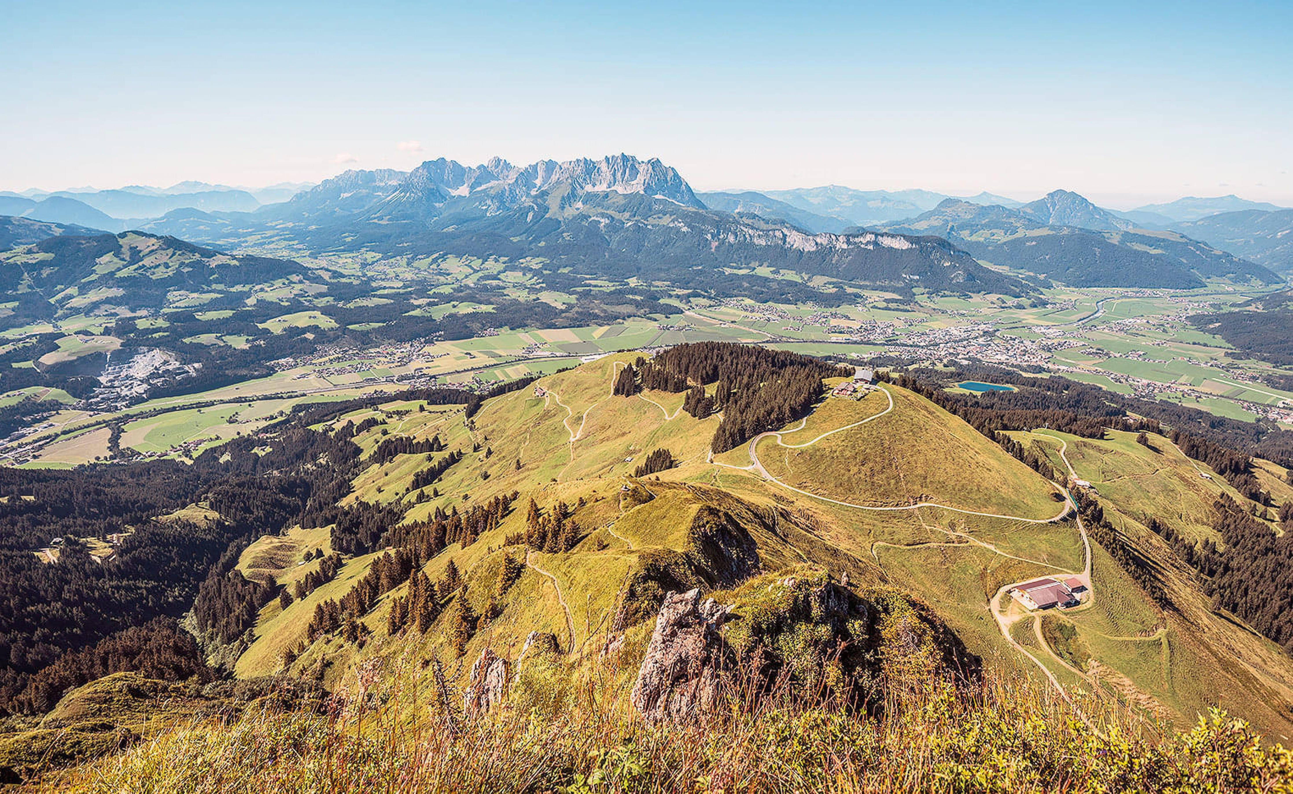 Weite Berglandschaft mit grünen Hügeln und Wanderwegen, im Hintergrund eine beeindruckende Gebirgskette unter klarem, blauem Himmel.