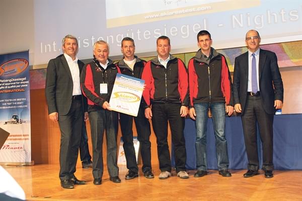 Sechs Männer stehen auf einer Bühne, einer hält eine Urkunde. Sie tragen Freizeit- oder Businesskleidung und lächeln in die Kamera.