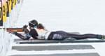 Sportlerin liegt im Schnee und zielt mit einem Gewehr bei einem Biathlon-Wettbewerb. Sie trägt Winterkleidung und einen Helm.