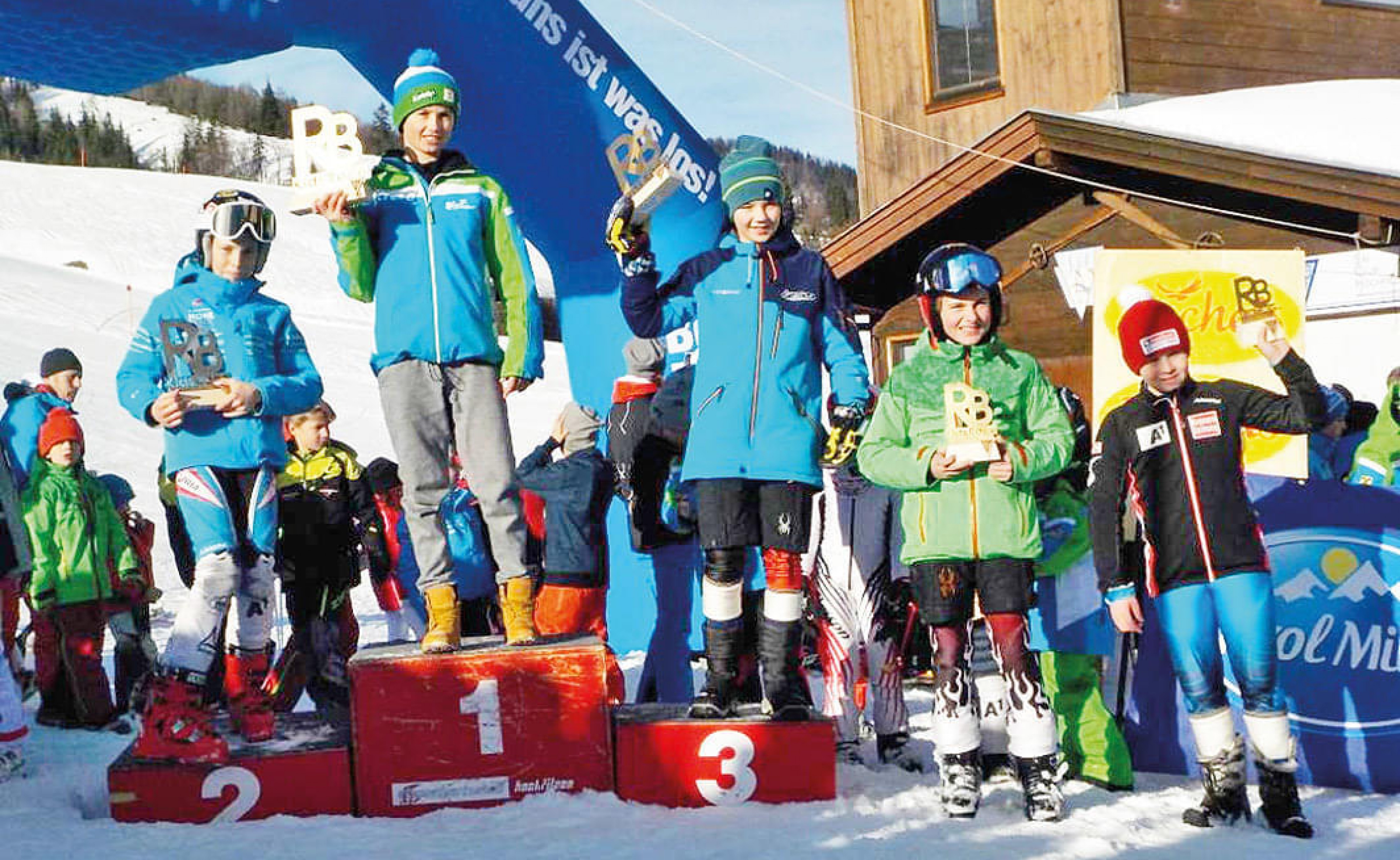 Kinder in Skibekleidung stehen auf einem Podium im Schnee mit Pokalen in der Hand, umgeben von anderen Teilnehmern vor einem Holzhäuschen und Schneehintergrund.