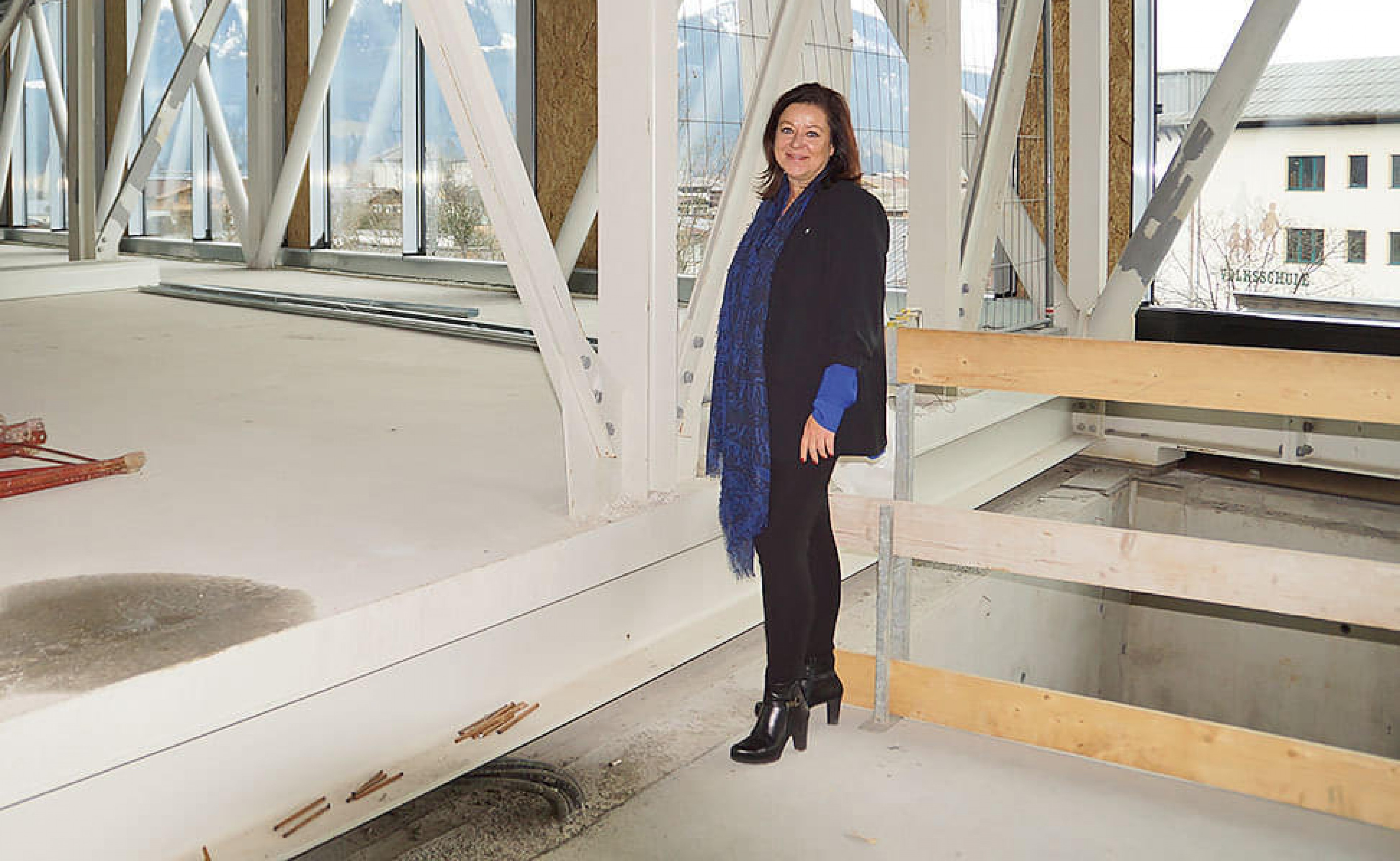 Frau steht in einem unfertigen Gebäudebereich mit Stahlträgern und Holzbalken, trägt schwarze Kleidung und einen blauen Schal.