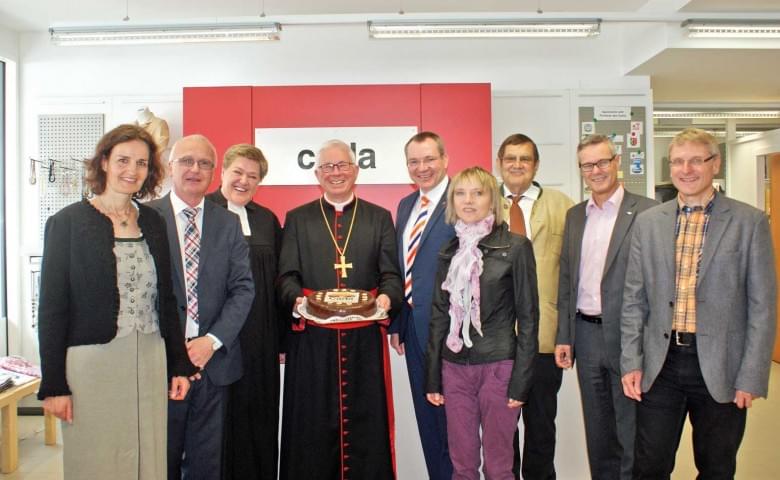 Gruppe von Personen in formeller Kleidung posiert mit einem Priester, der eine Torte trägt, vor einer roten Wand mit der Aufschrift "carla".