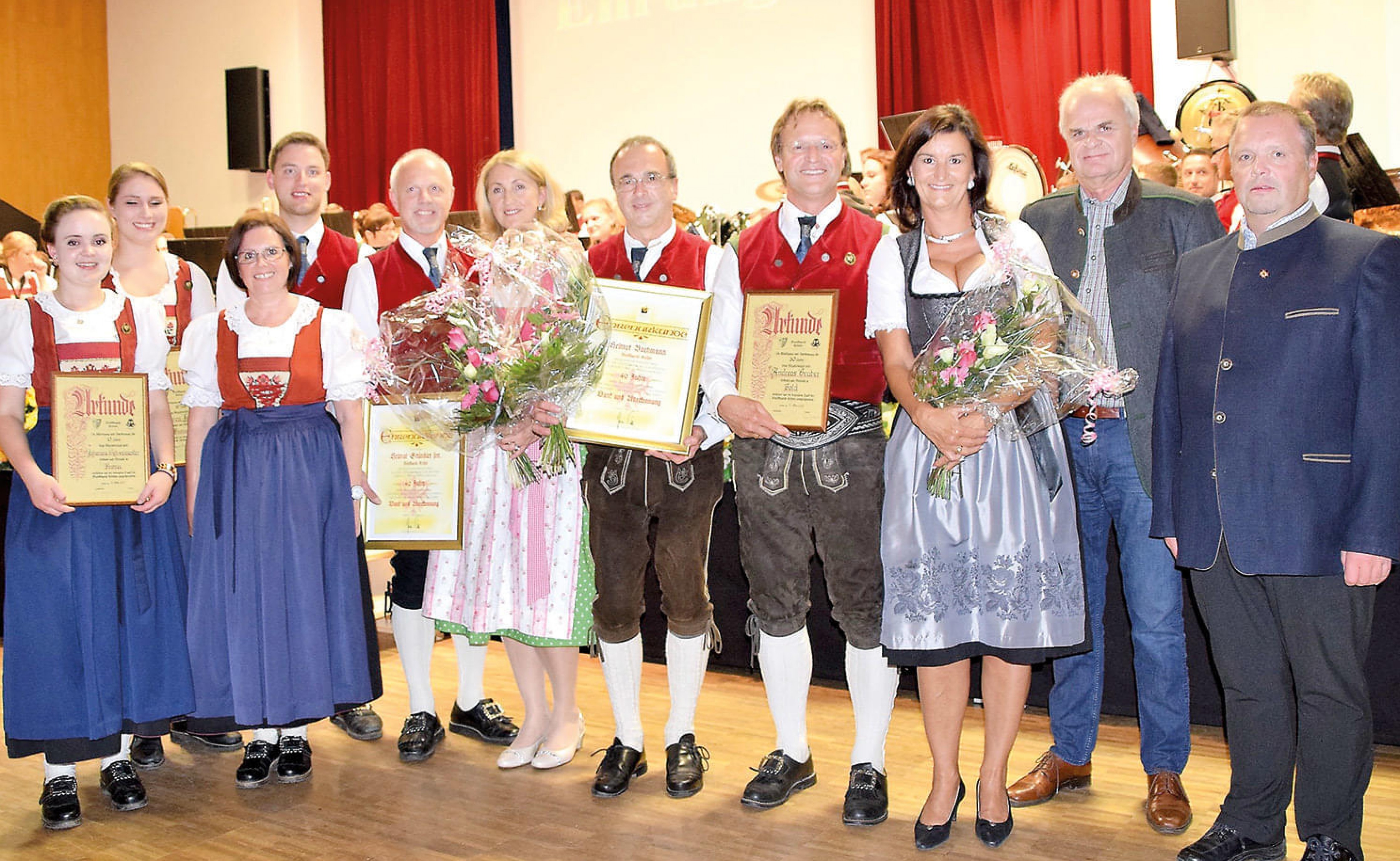 Eine Gruppe von Menschen in traditioneller Tracht mit Blumensträußen und Urkunden, posierend für ein Foto in einem festlichen Saal.