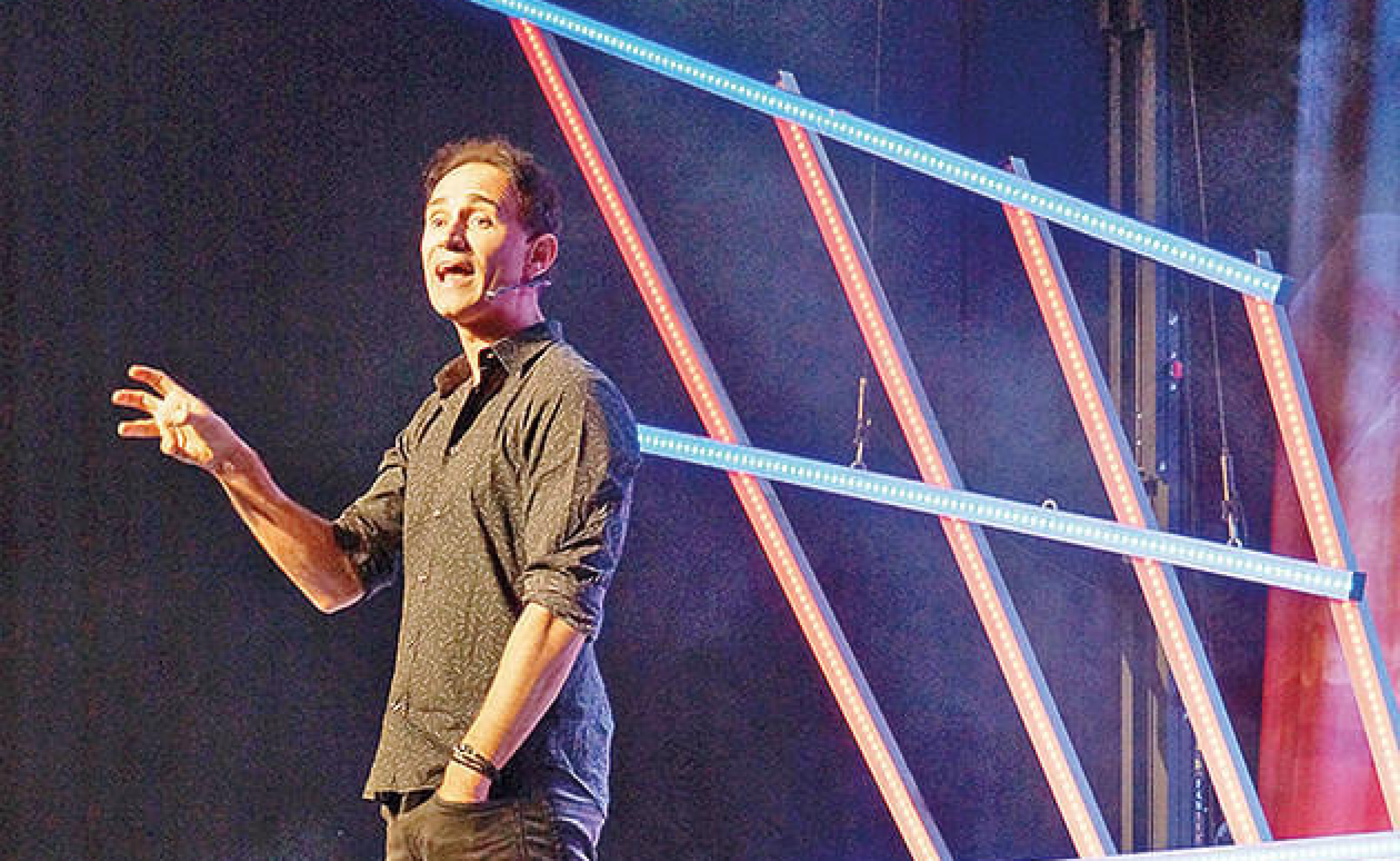 Ein Mann in dunklem Hemd spricht gestikulierend auf einer Bühne, hinter ihm leuchten schräge LED-Streifen in Blau und Rot. Die Atmosphäre ist dynamisch.