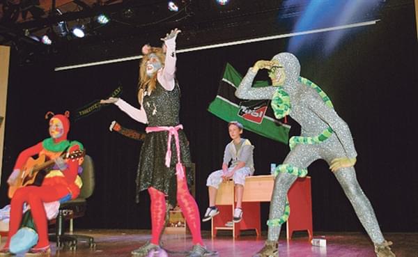 Künstler in bunten, schrillen Kostümen performen lebhaft auf einer Bühne vor einem schwarzen Vorhang mit Fahne im Hintergrund.