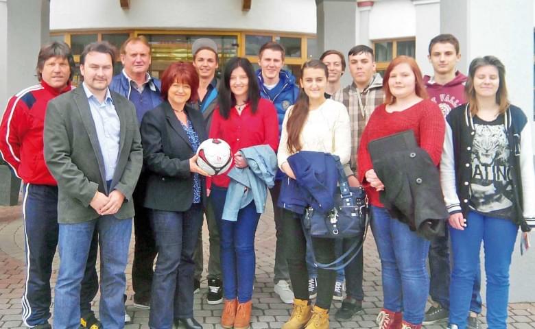 Eine Gruppe von Menschen posiert lächelnd für ein Foto vor einem Gebäude. Eine Person in der Mitte hält einen Fußball. Sie tragen lässige Kleidung.