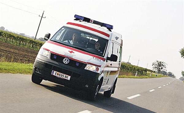 Ein Krankenwagen fährt bei Tageslicht auf einer Landstraße entlang, umgeben von Feldern und Bäumen, mit Personen im Inneren.