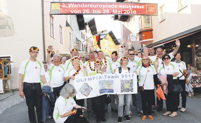 Eine Gruppe von Menschen in weißen und grünen T-Shirts posiert fröhlich auf einer Straße unter einem Banner für die Wandereuropiade im Mai 2016.