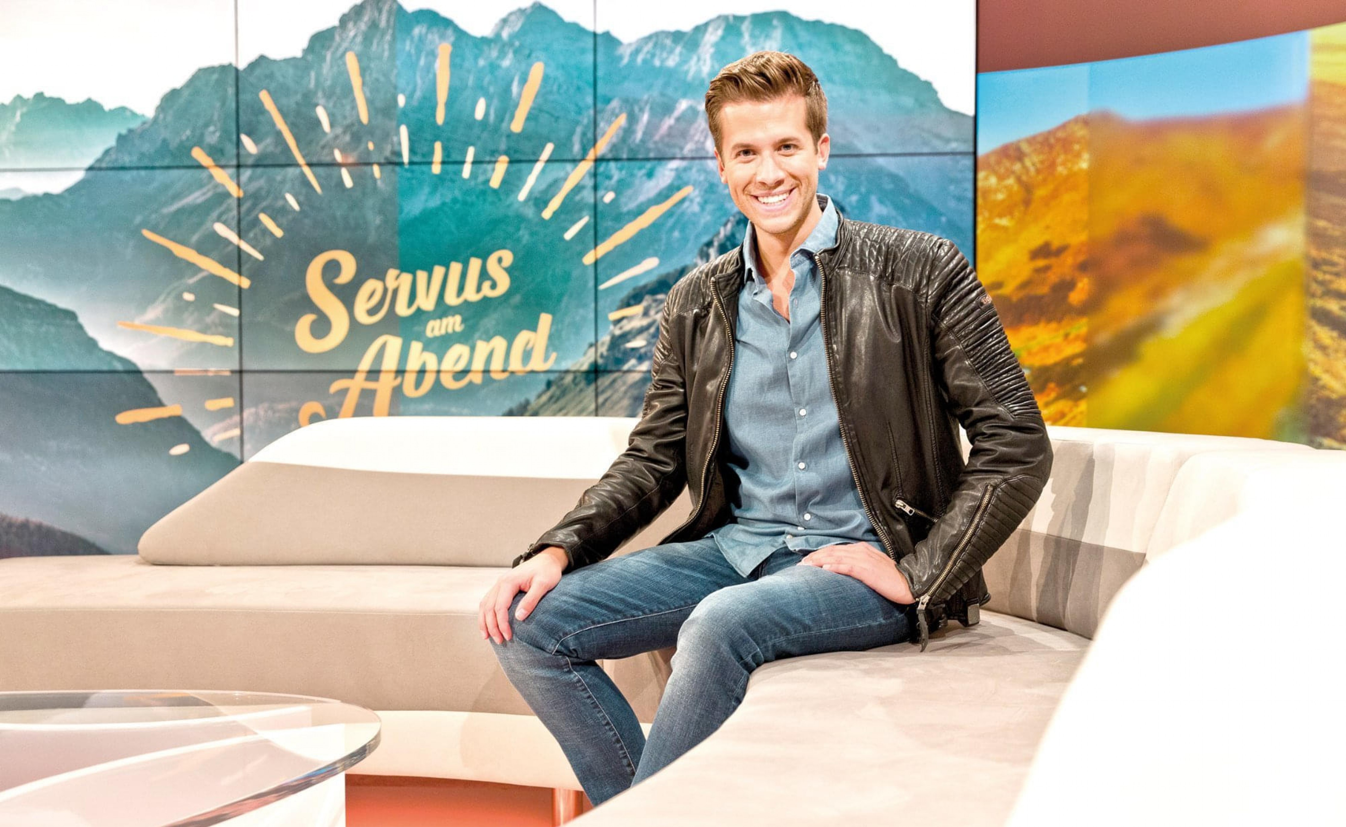 Ein lächelnder Moderator sitzt auf einem Sofa vor einem Bildschirm, der "Servus am Abend" zeigt, mit Bergen im Hintergrund. Er trägt Freizeitkleidung.