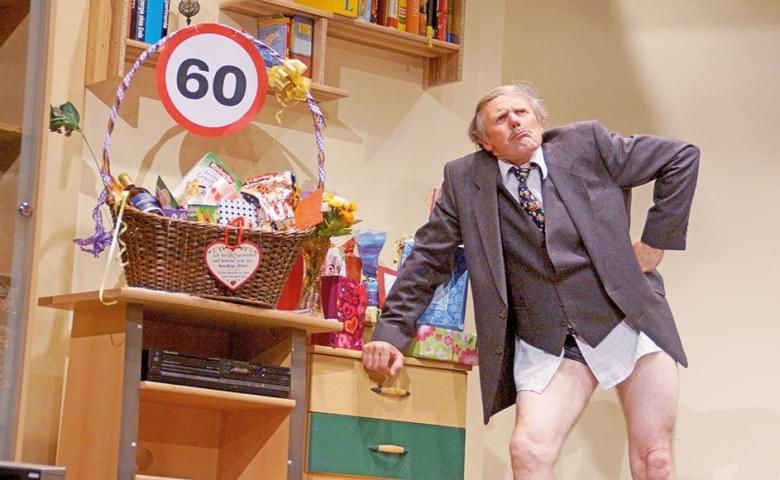 Ein älterer Mann in Anzug steht mit entblößten Beinen neben einem Geschenkkorb und einem Schild mit der Zahl 60 in einem Wohnzimmer voller Geschenke.