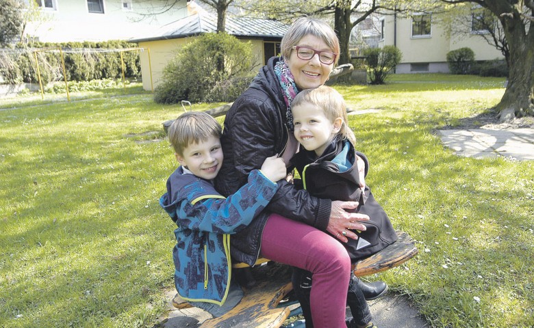 Eine lächelnde Person sitzt auf einer Bank im Park und hält zwei Kinder im Arm. Die Kinder tragen Jacken und schauen fröhlich. Im Hintergrund sind Bäume und ein Gebäude.