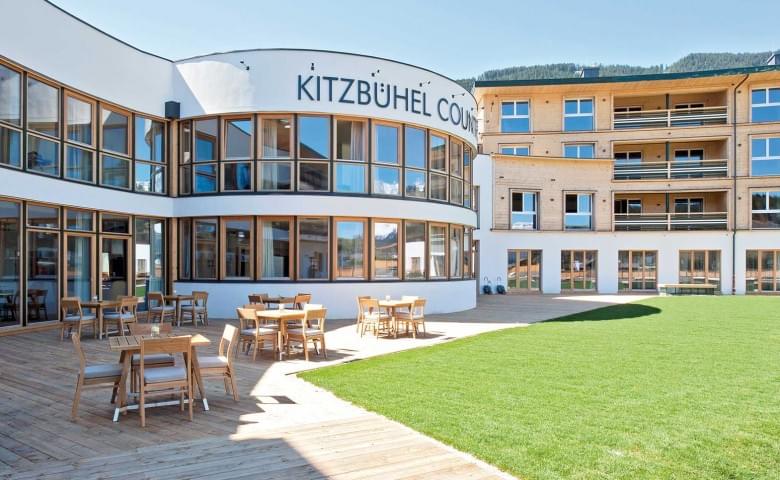 Ein modernes Hotelgebäude in Holzbauweise mit einem runden Eingangsbereich und großen Fenstern. Davor stehen Tische und Stühle auf einer Terrasse.