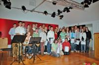 Eine Gruppe von Menschen steht auf einer Bühne. Einige halten Urkunden. Im Vordergrund ein Musiker mit einem Instrument. Der Hintergrund ist rot beleuchtet.