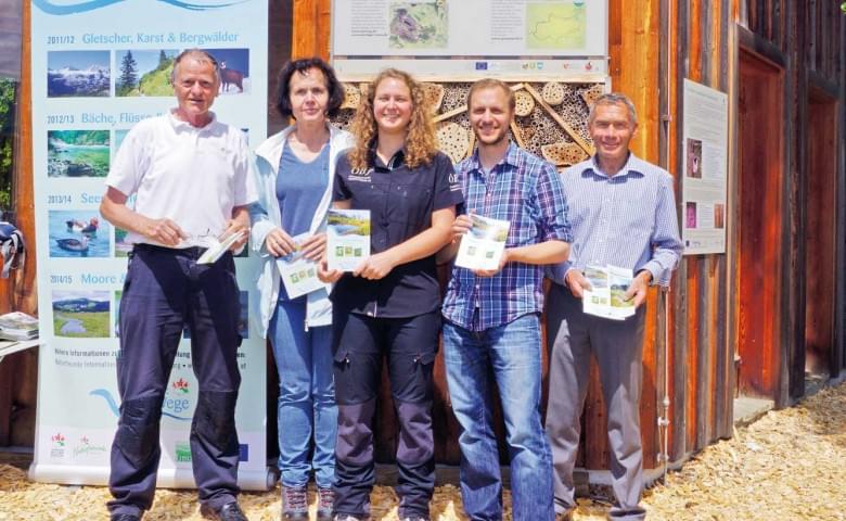Eine Gruppe von fünf Menschen steht vor einem Informationsstand aus Holz und hält Broschüren in der Hand. Im Hintergrund sind Texttafeln und Plakate zu sehen.