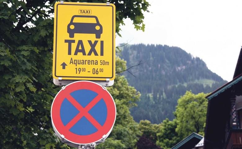 Gelbes Taxi-Schild zeigt den Weg zur Aquarena an, daneben ein Halteverbotsschild vor einer Bergkulisse und Bäumen.