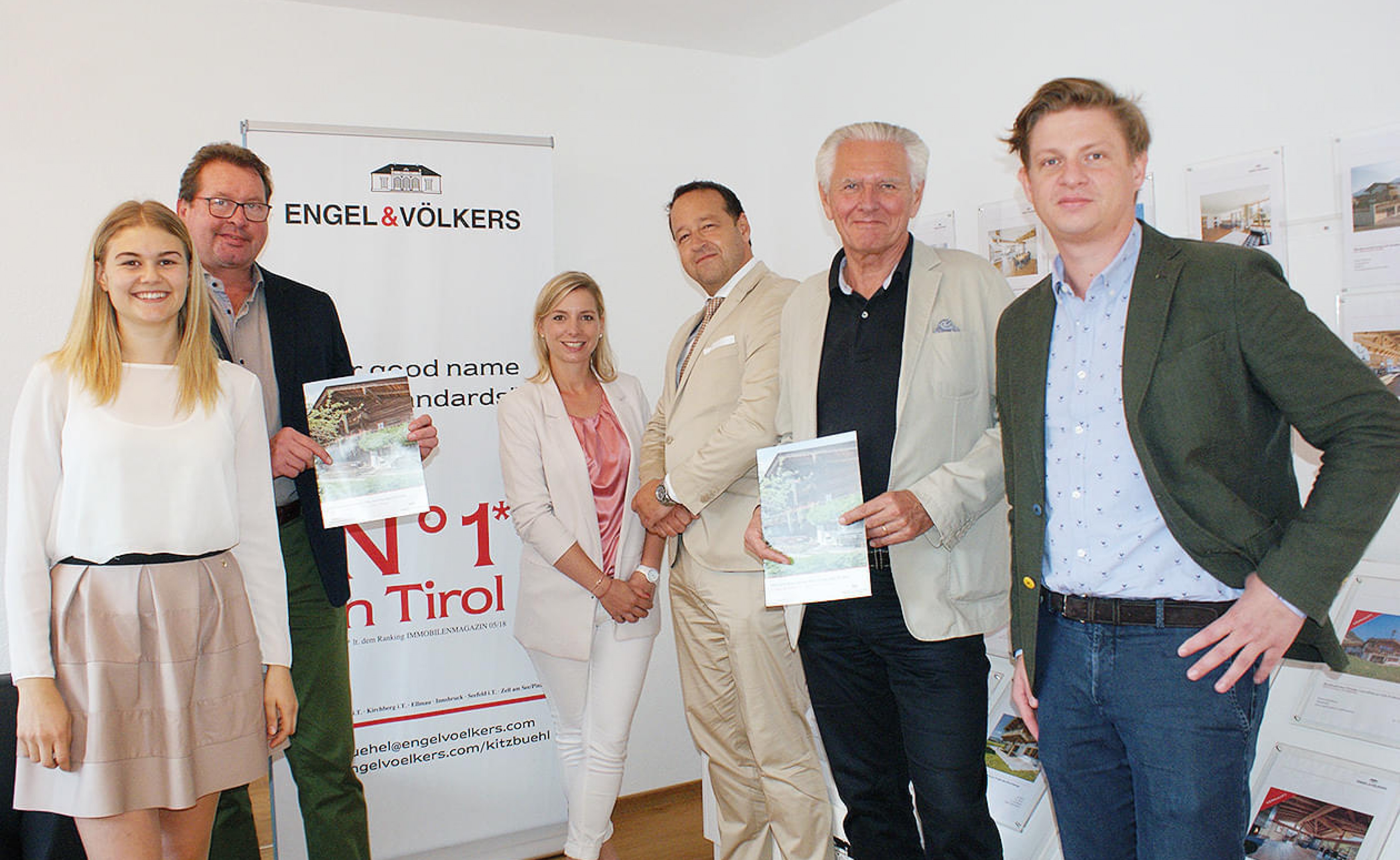 Sechs Personen stehen in einem Büro vor einem Roll-up von Engel & Völkers und halten Broschüren. Sie lächeln in die Kamera und tragen formelle Kleidung.