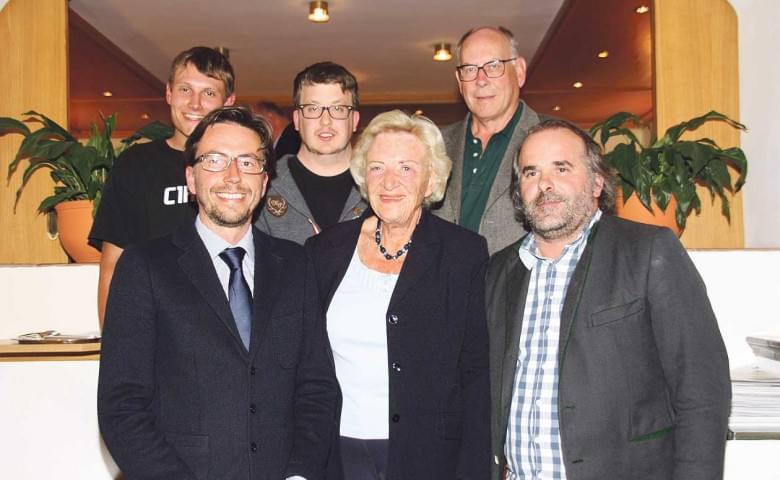 Eine Gruppe von sechs Menschen in formeller Kleidung posiert lächelnd für ein Foto in einem Innenraum, umgeben von Pflanzen und Holzvertäfelung.