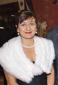 Frau in eleganter Kleidung mit weißem Pelz und Perlenkette, lächelt in die Kamera. Im Hintergrund sind unscharfe Personen erkennbar.