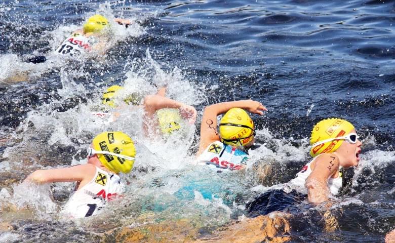 Gruppe von Triathleten mit gelben Badekappen und Sonnenbrillen schwimmen in offenem Wasser während eines Wettbewerbs.
