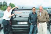 Drei Männer stehen vor einem Auto mit einer Werbebotschaft auf der Heckscheibe, in einem Parkplatz umgeben von Bäumen und Bergen im Hintergrund.