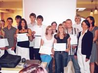 Gruppe von Menschen, teils mit Zertifikaten, posiert lächelnd vor einer Leinwand in einem Innenraum mit warmer Beleuchtung.