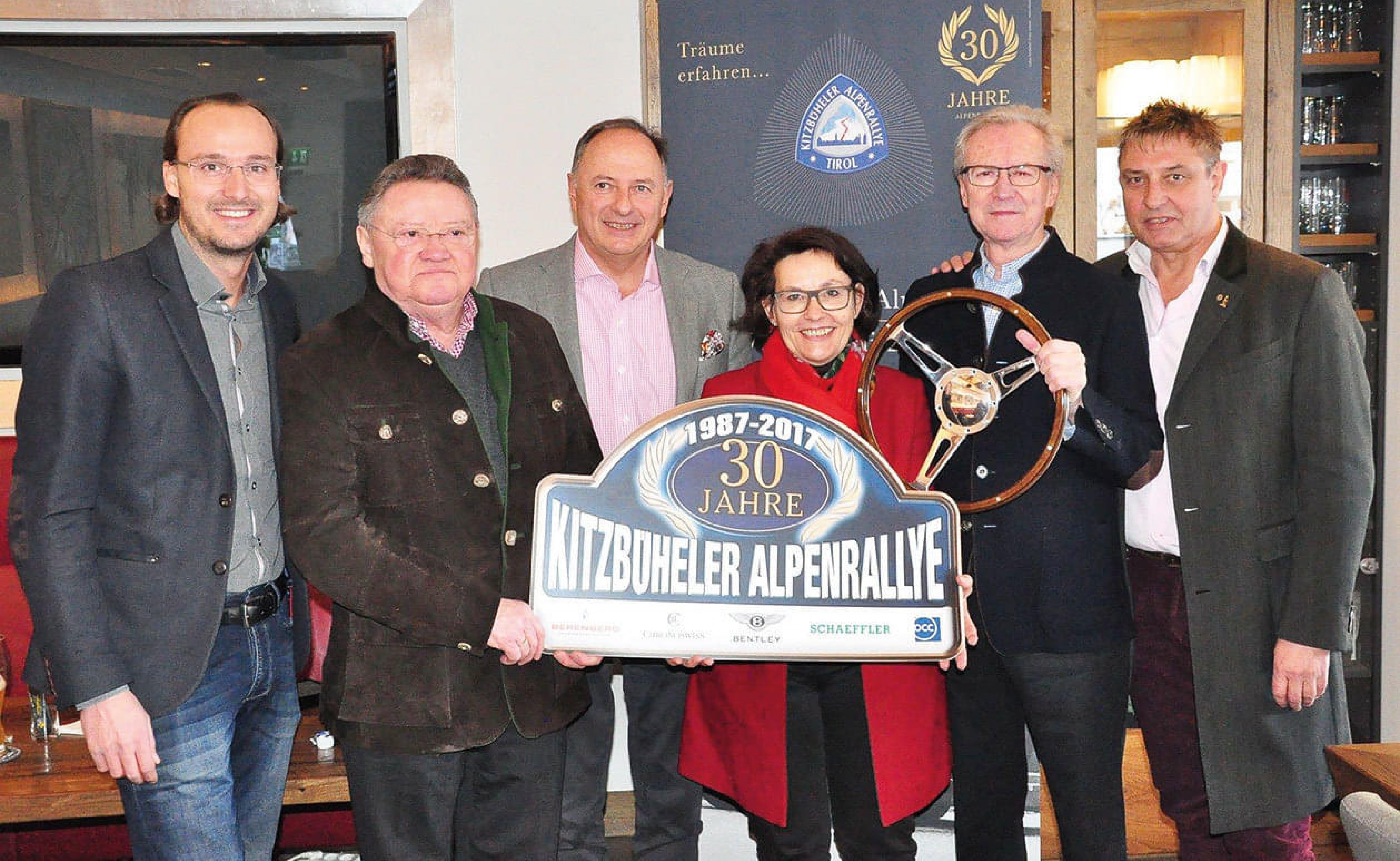 Gruppenfoto von sieben Personen, die ein Jubiläumsschild und ein Lenkrad halten, um das 30-jährige Bestehen der Kitzbüheler Alpenrallye zu feiern.