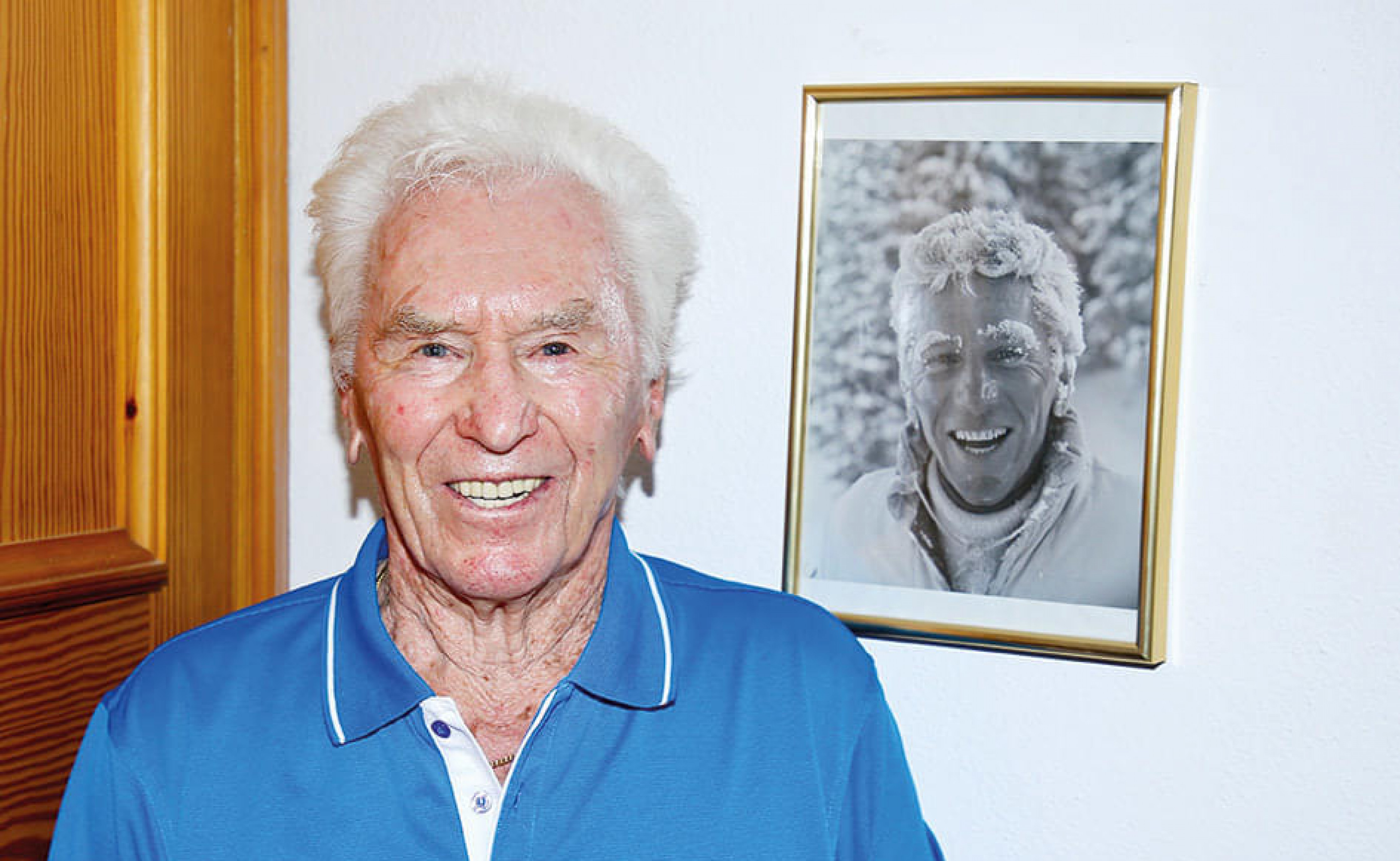 Ein älterer Mann mit weißem Haar lächelt in die Kamera. Im Hintergrund hängt ein gerahmtes Schwarz-Weiß-Foto einer lächelnden Person an der Wand.