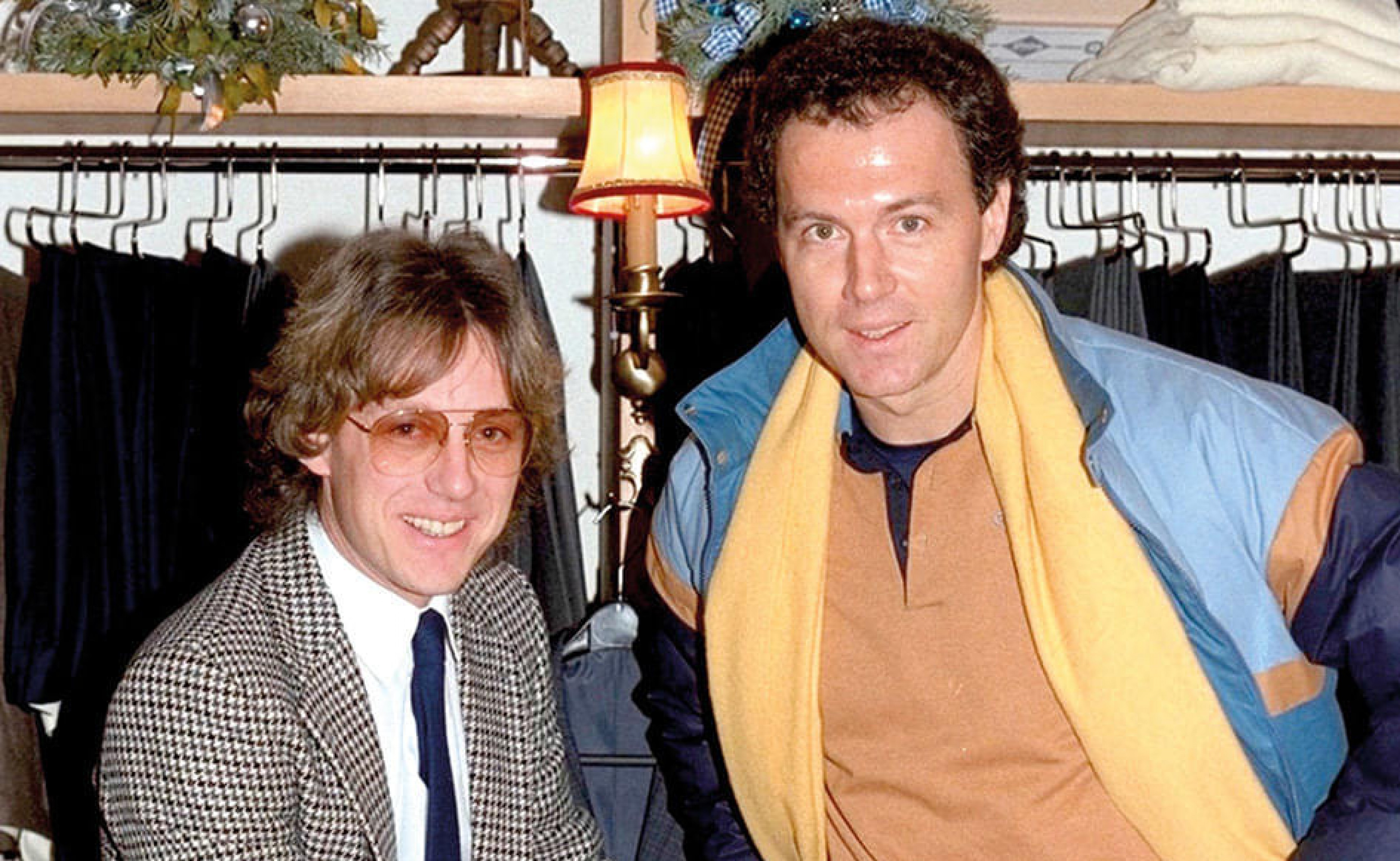 Zwei Männer in einem Geschäft, einer trägt eine Karojacke und Brille, der andere eine blaue Jacke mit gelbem Schal. Im Hintergrund sind Kleider und eine Lampe zu sehen.