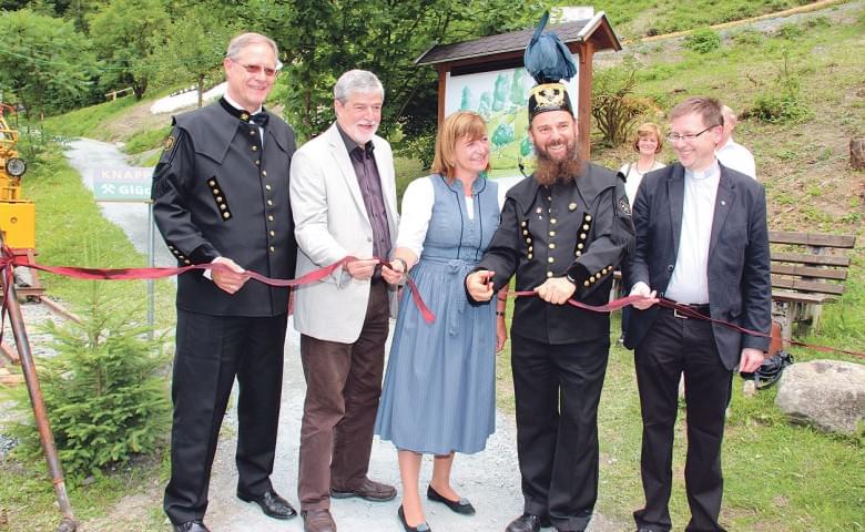 Eine feierliche Eröffnung im Freien mit sechs Personen, die ein rotes Band durchschneiden. Zwei tragen festliche Hüte und traditionelle Bergmannskleidung.