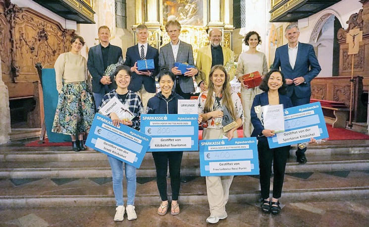 Eine Gruppe von Menschen steht in einem festlichen Raum, einige halten große Schecks mit Geldbeträgen. Im Hintergrund sind kunstvolle Dekorationen zu sehen.