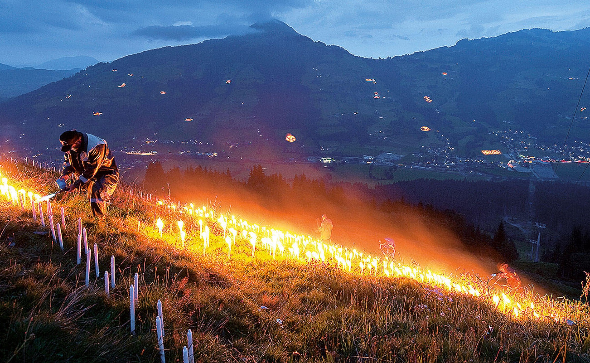 Ein Mann zündet in einer hügeligen Landschaft Kerzen oder Fackeln an, während der Himmel in der Abenddämmerung blau ist. Im Hintergrund sind Berge zu sehen.