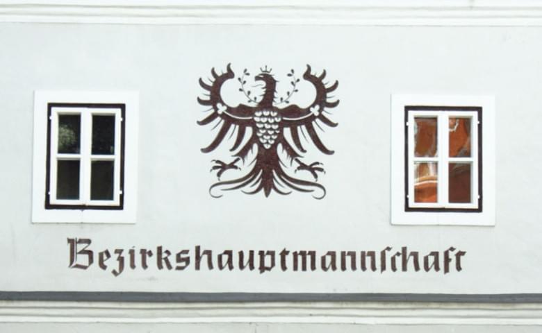 Historisches Gebäude mit zwei weißen Fenstern und einem Wappen eines Adlers, darunter das Wort 'Bezirkshauptmannschaft' auf heller Fassade.